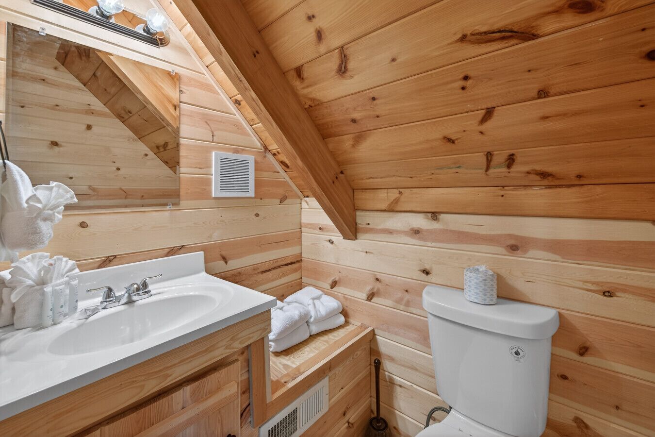 A wooden cabin bathroom with a sink, mirror, toilet, and a vent.