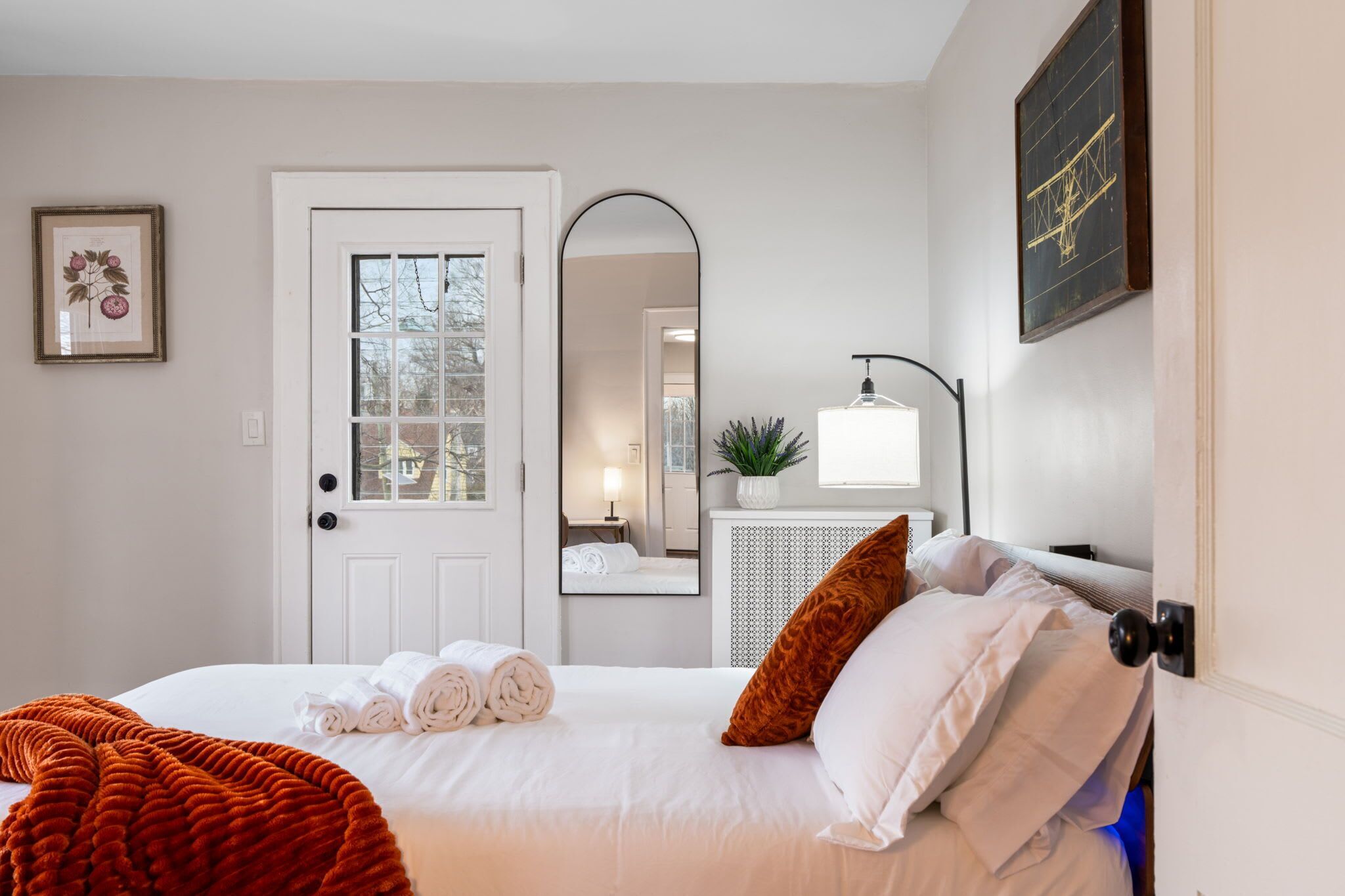 A hotel room with a bed, a door, a mirror, a plant, and a wall-mounted light fixture.