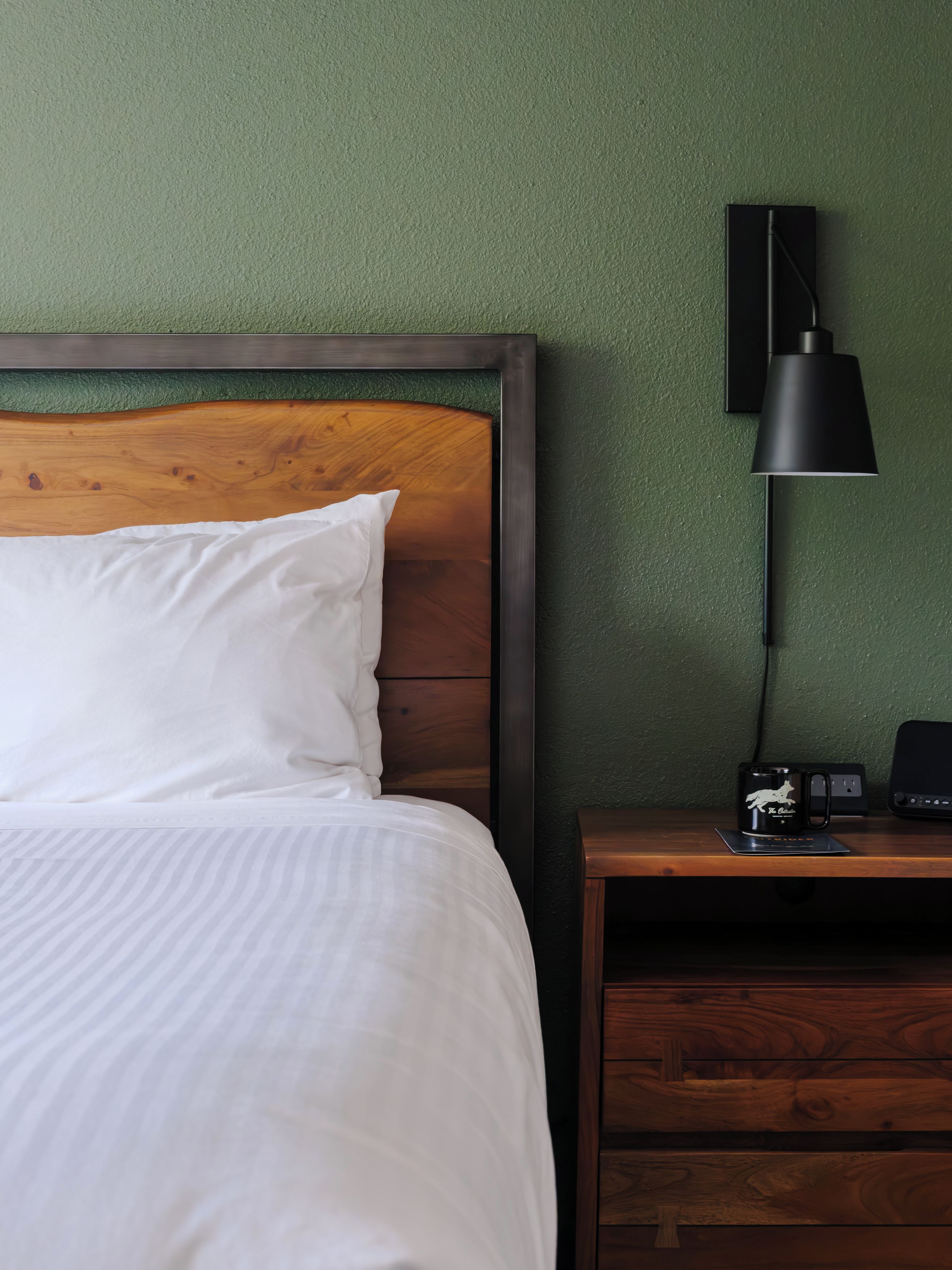 Una cama bien hecha con cabecera de madera, una mesita de noche con lámpara y un enchufe en la pared.