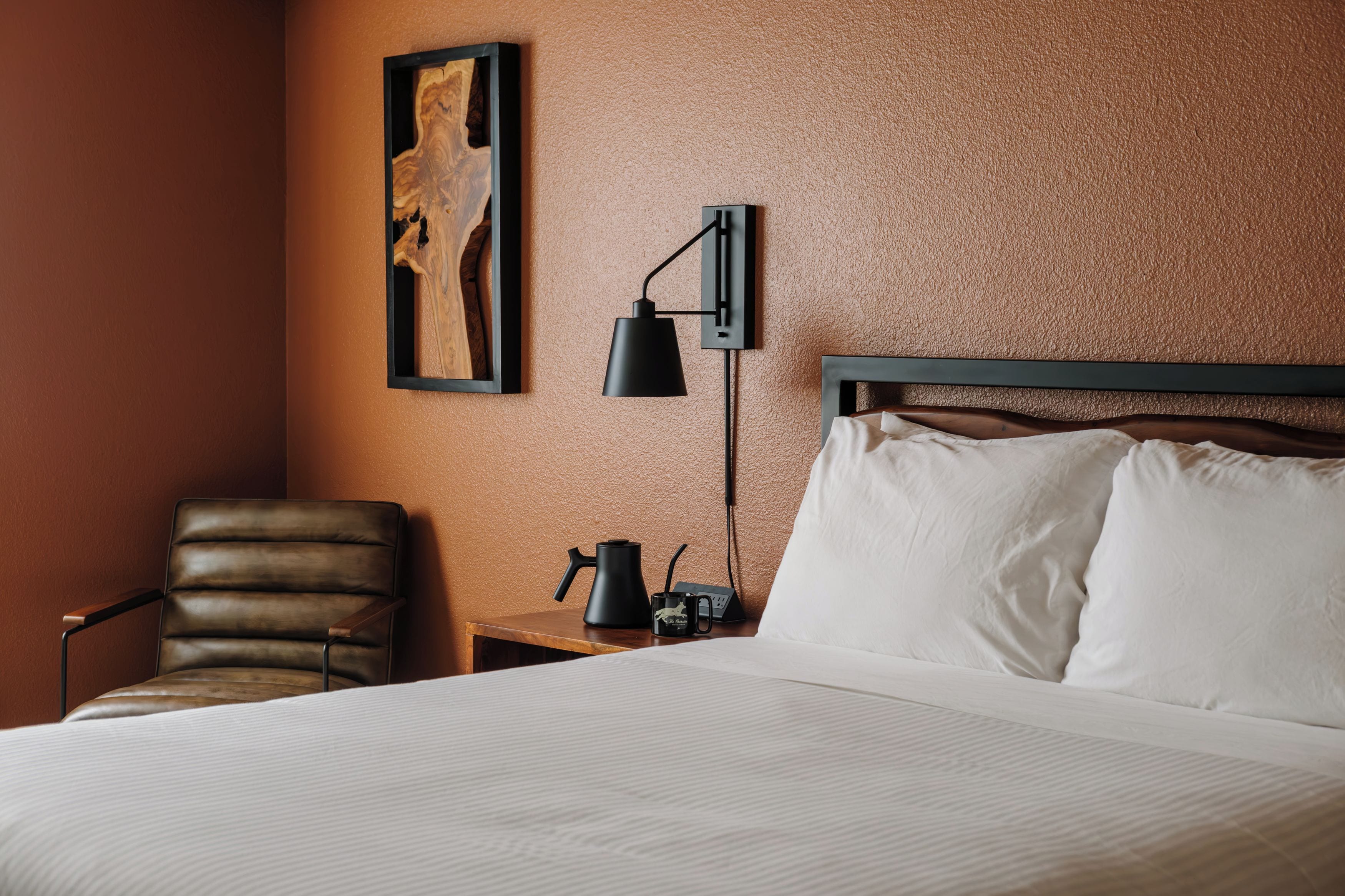 A hotel room with a bed, a chair, a nightstand with a lamp, and a painting on the wall.