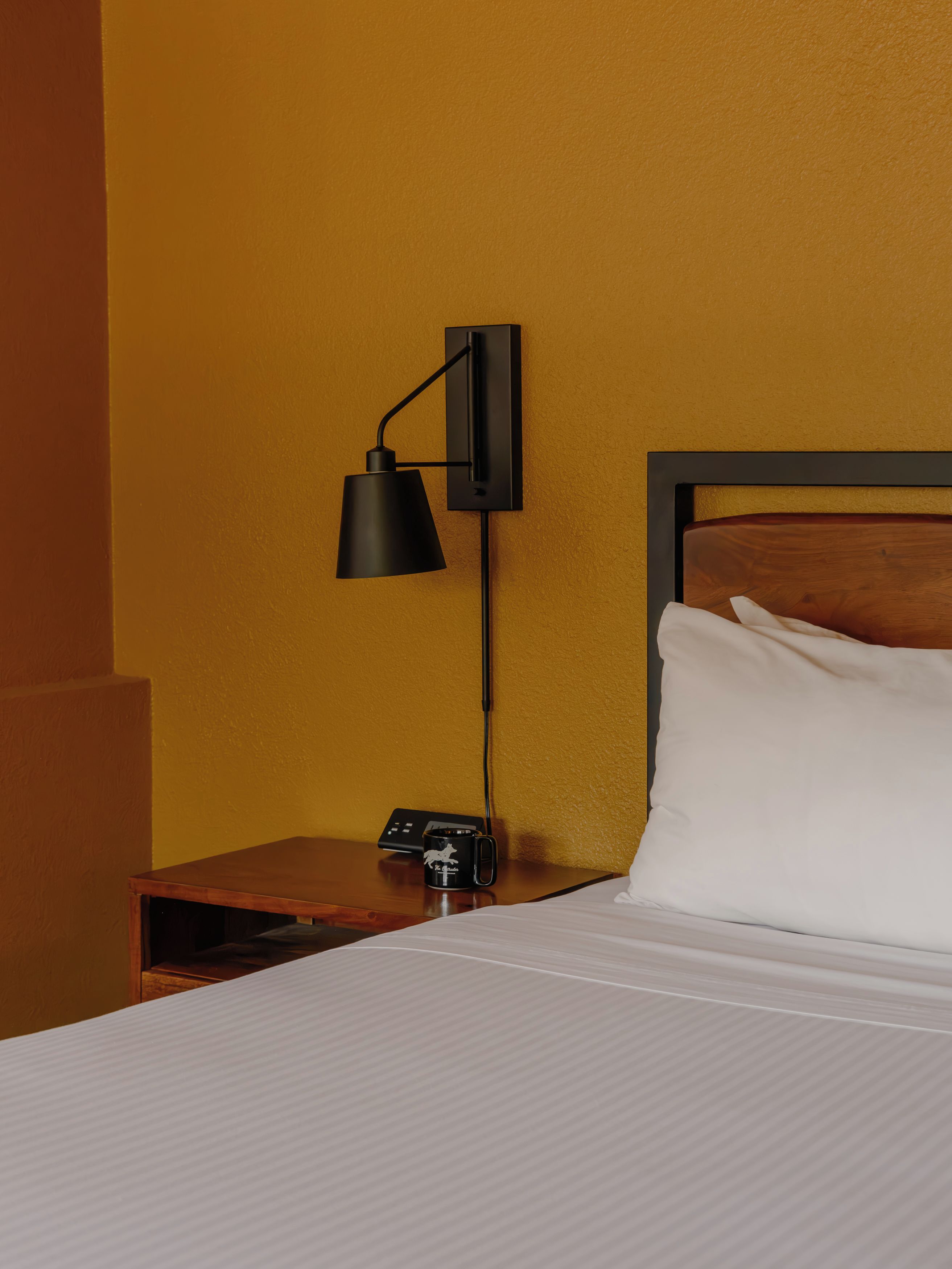Una habitación de hotel con una cama, una mesita de noche, una lámpara de escritorio negra, un control remoto y una taza negra con un diseño blanco.