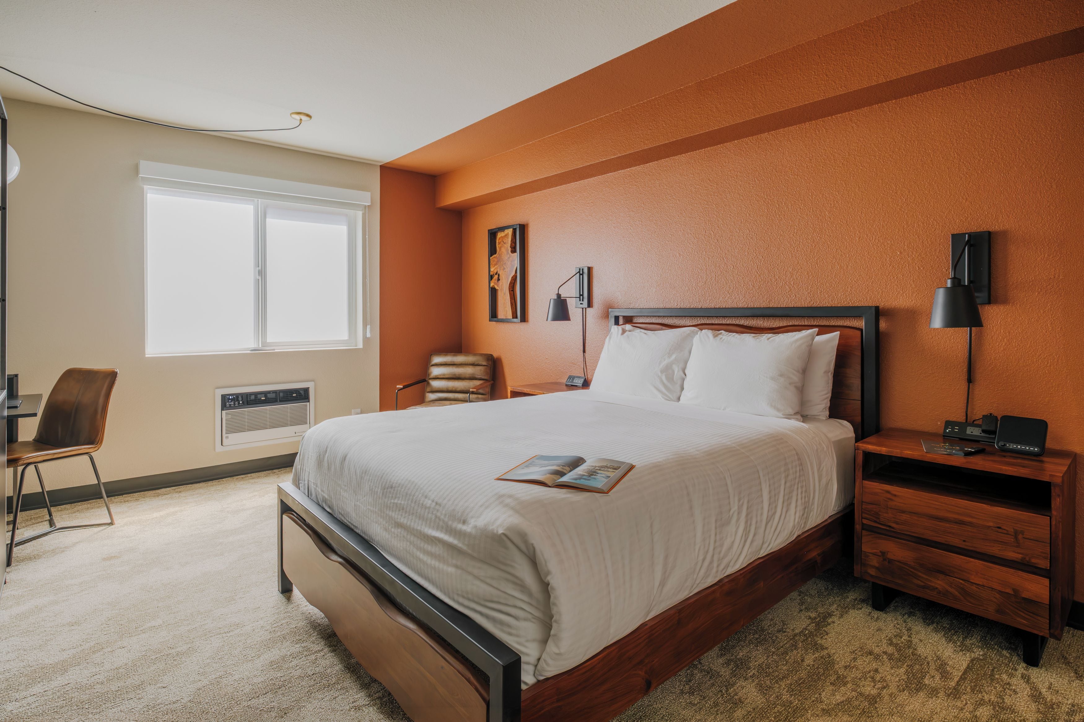 A hotel room with a large bed, a desk, a chair, a bedside table with a lamp, and a wall-mounted artwork.