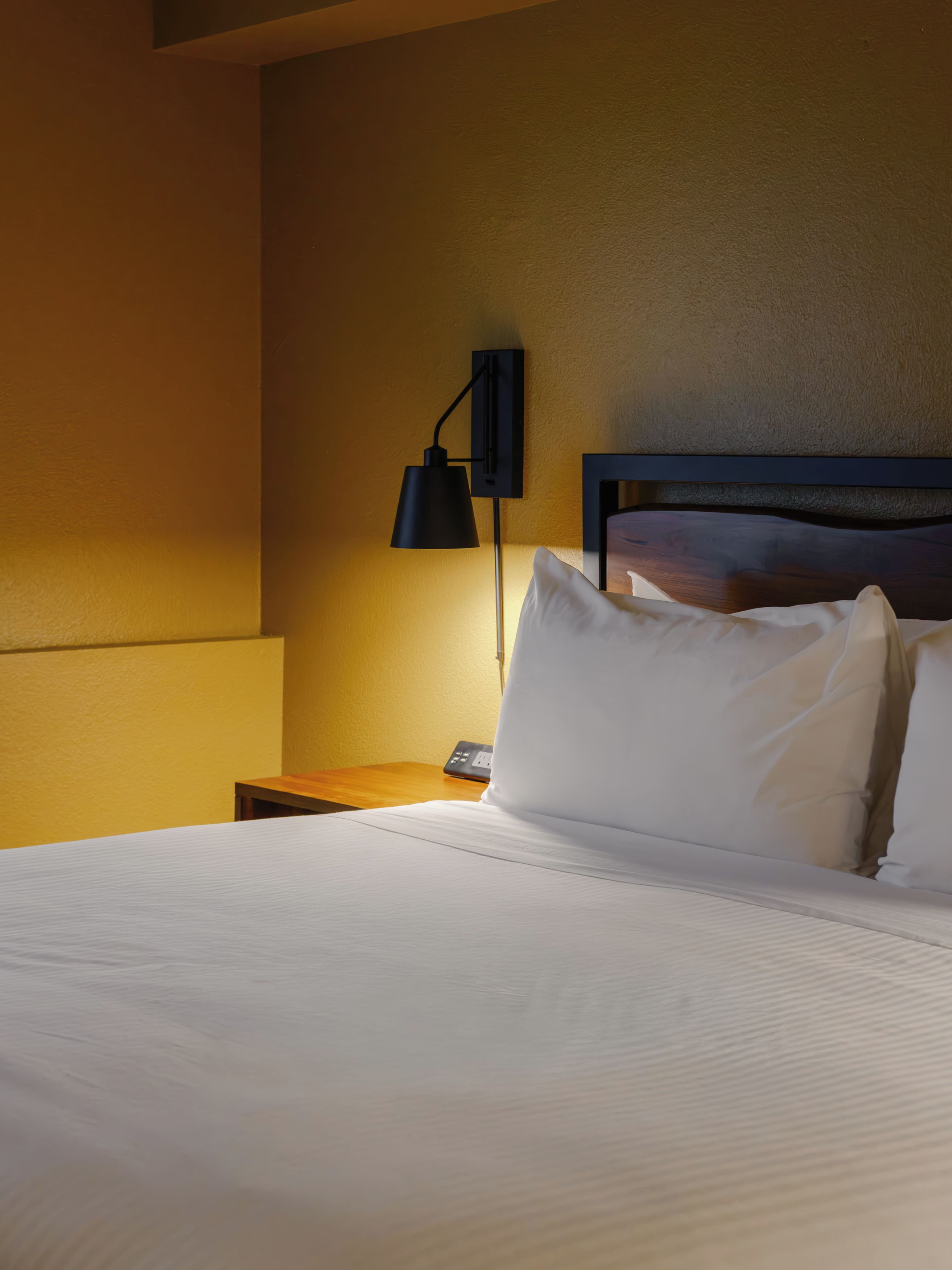 A hotel room with a neatly made bed, a bedside table, a lamp, and a wall-mounted light fixture.