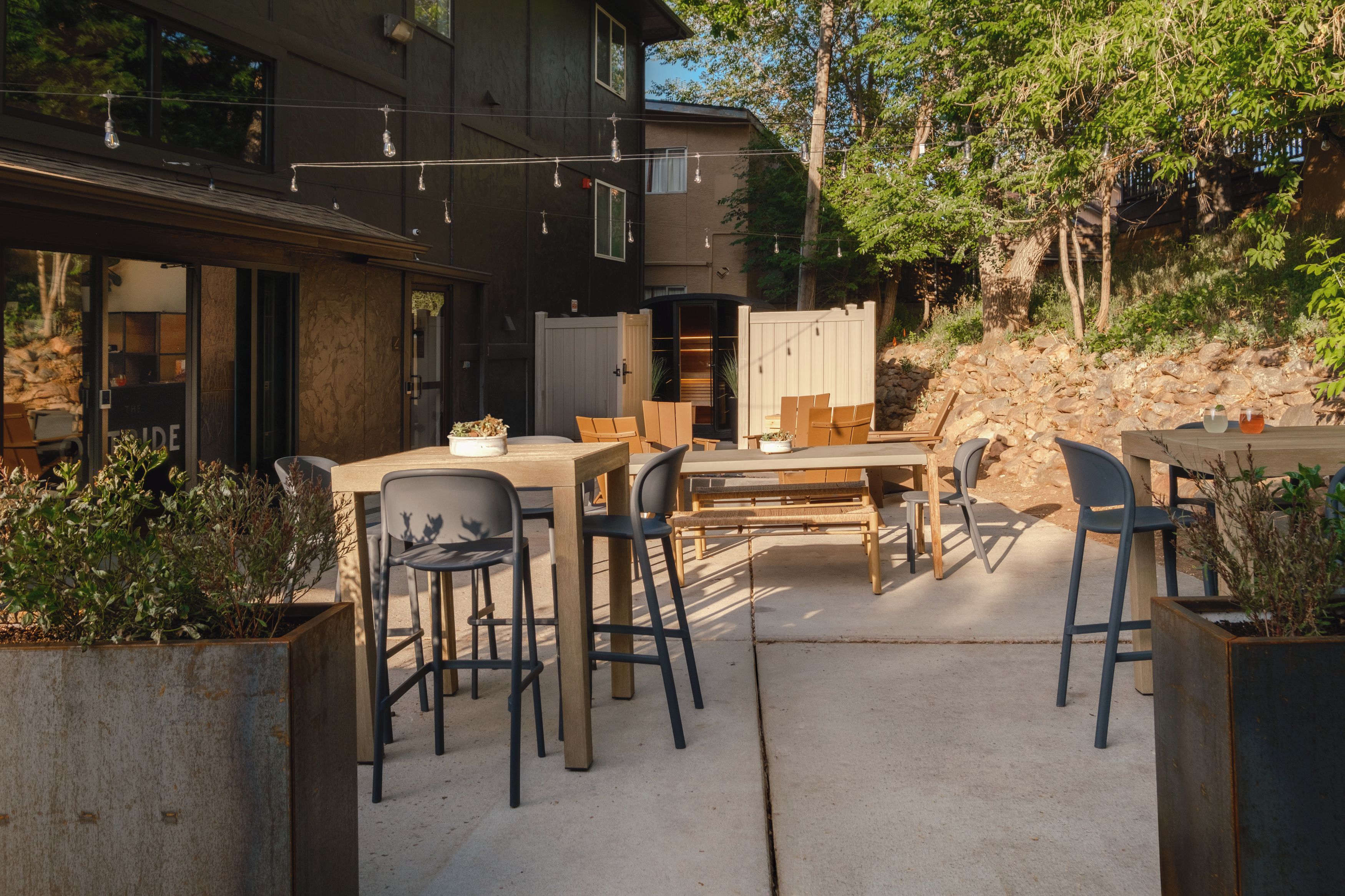 An outdoor patio area with a dining table and chairs, surrounded by planters and a building with large windows.