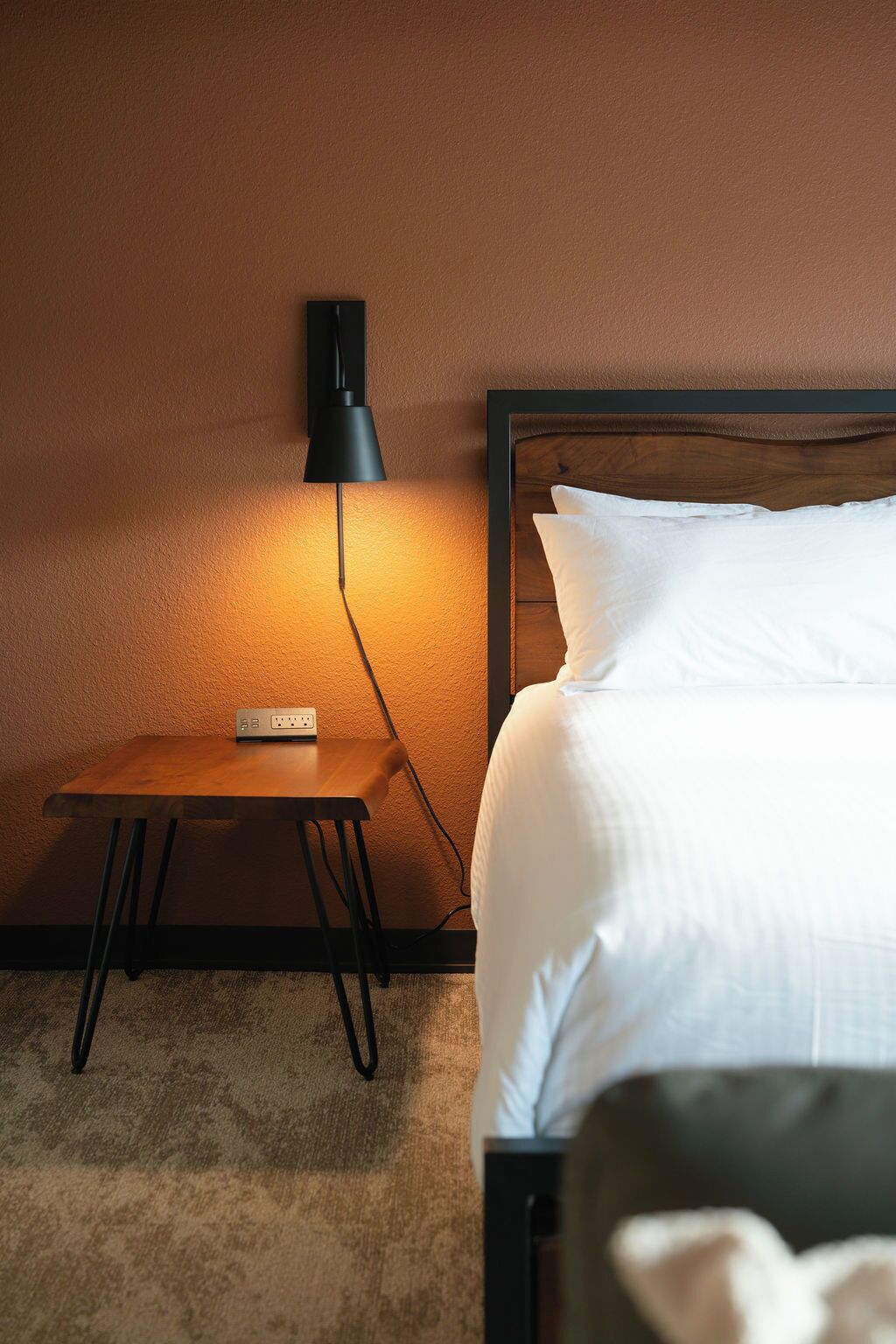 Une chambre d’hôtel avec une table de chevet en bois, une lampe fixée au mur, un lit avec une literie blanche et un sol recouvert de moquette.