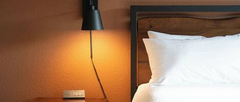 A hotel room with a wooden bedside table, a wall-mounted lamp, a bed with white bedding, and a carpeted floor.