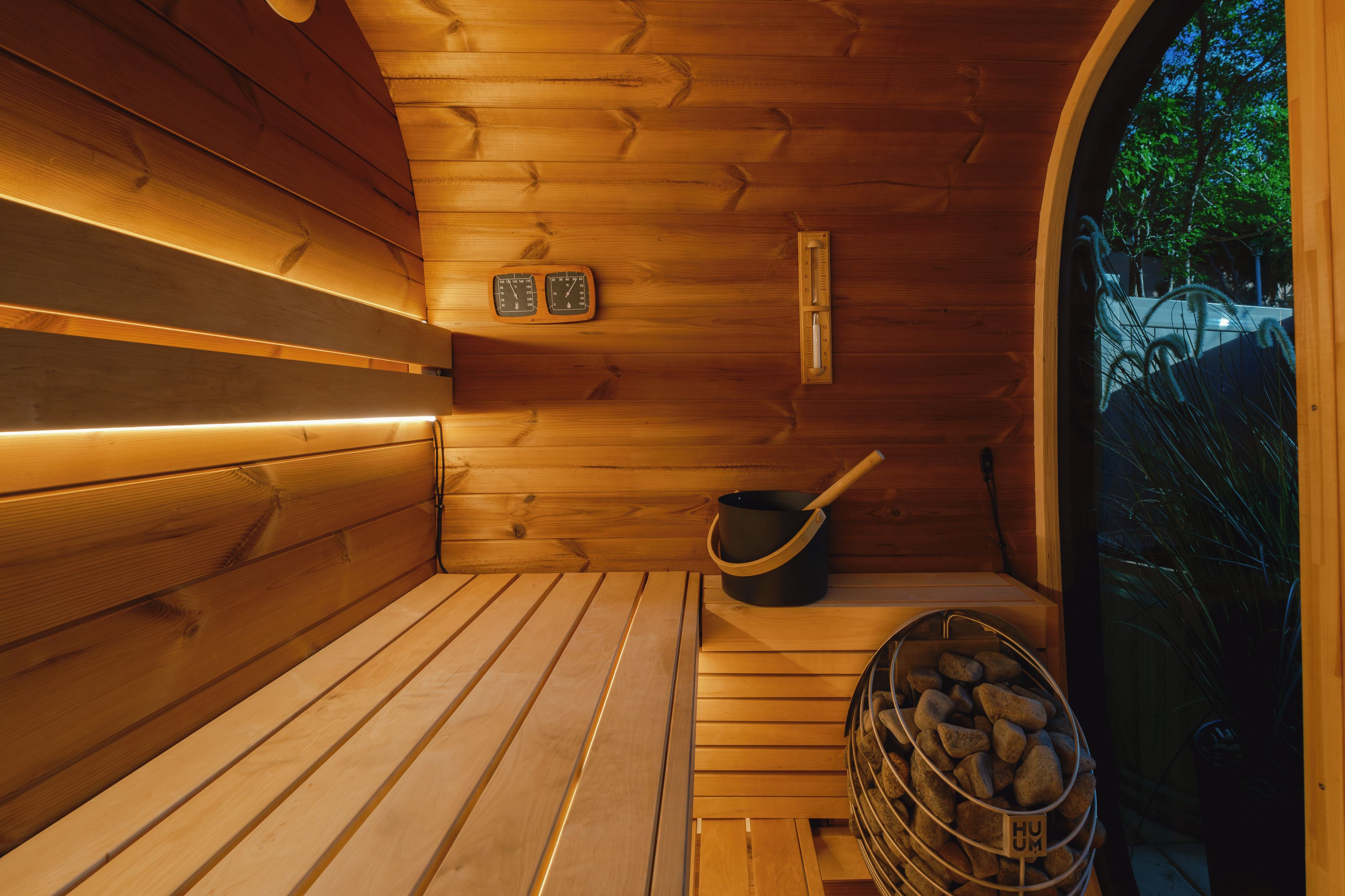 Una sauna con paredes de madera, un banco, un termómetro, un cubo y un recipiente con piedras.