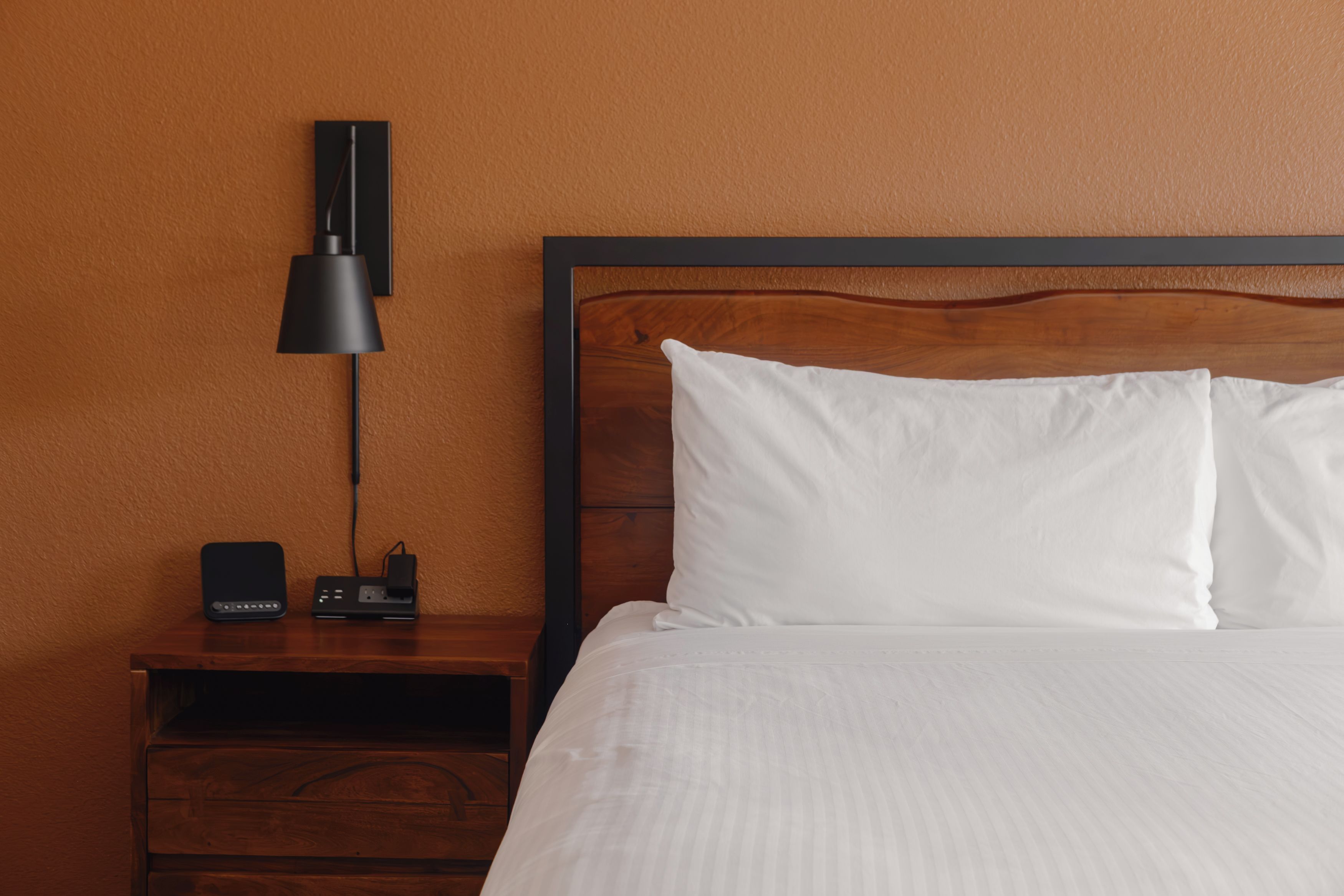 A hotel room with a wooden nightstand, a black lamp, a power strip, and a bed with white bedding.
