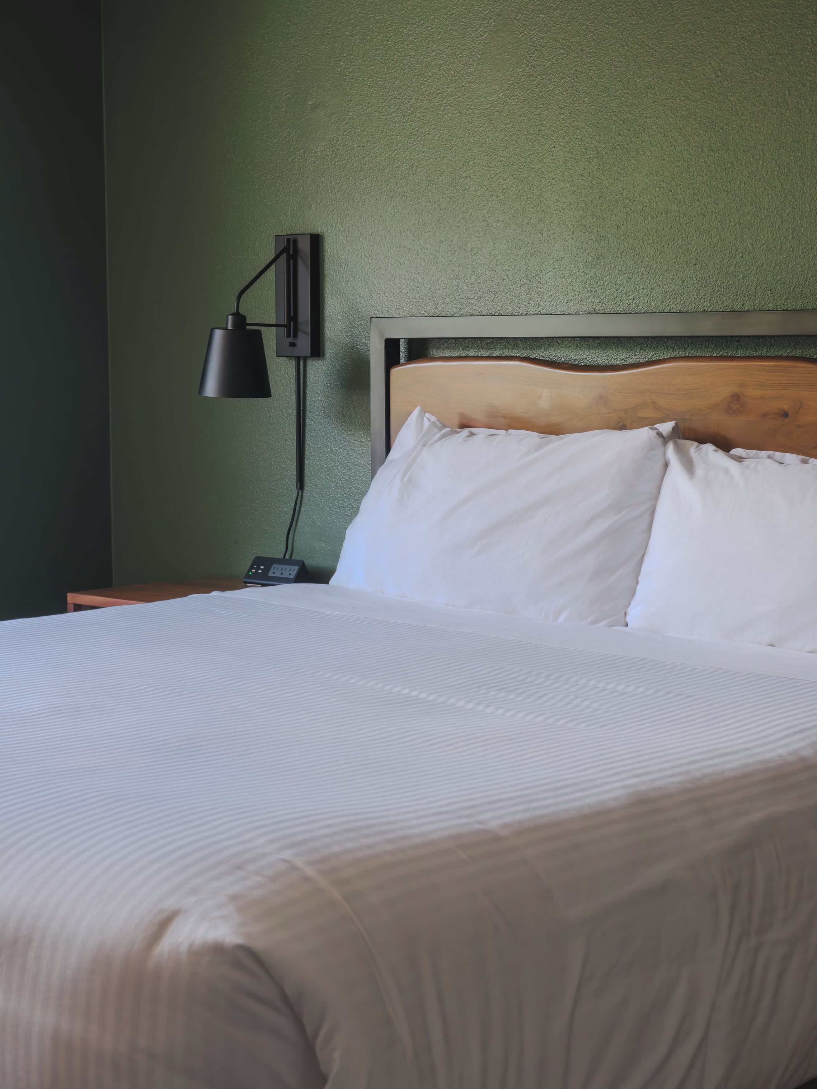 Een net opgemaakt bed met witte lakens, een houten hoofdbord en een wandlamp.