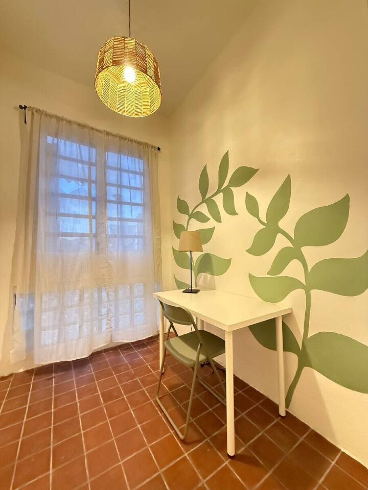Une pièce avec une table blanche, une chaise, une lampe, une fenêtre avec des rideaux légers, un mur orné d’un motif de feuilles et un luminaire suspendu en matière tressée.