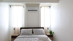 Un dormitorio con una cama, dos mesitas de noche, una planta en maceta y un equipo de aire acondicionado.