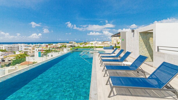 A rooftop infinity pool with clear blue water, surrounded by lounge chairs and a view of the ocean and cityscape. - Vibrante y Moderno Estudio Cerca de Quinta Avenida (Playa del Carmen)