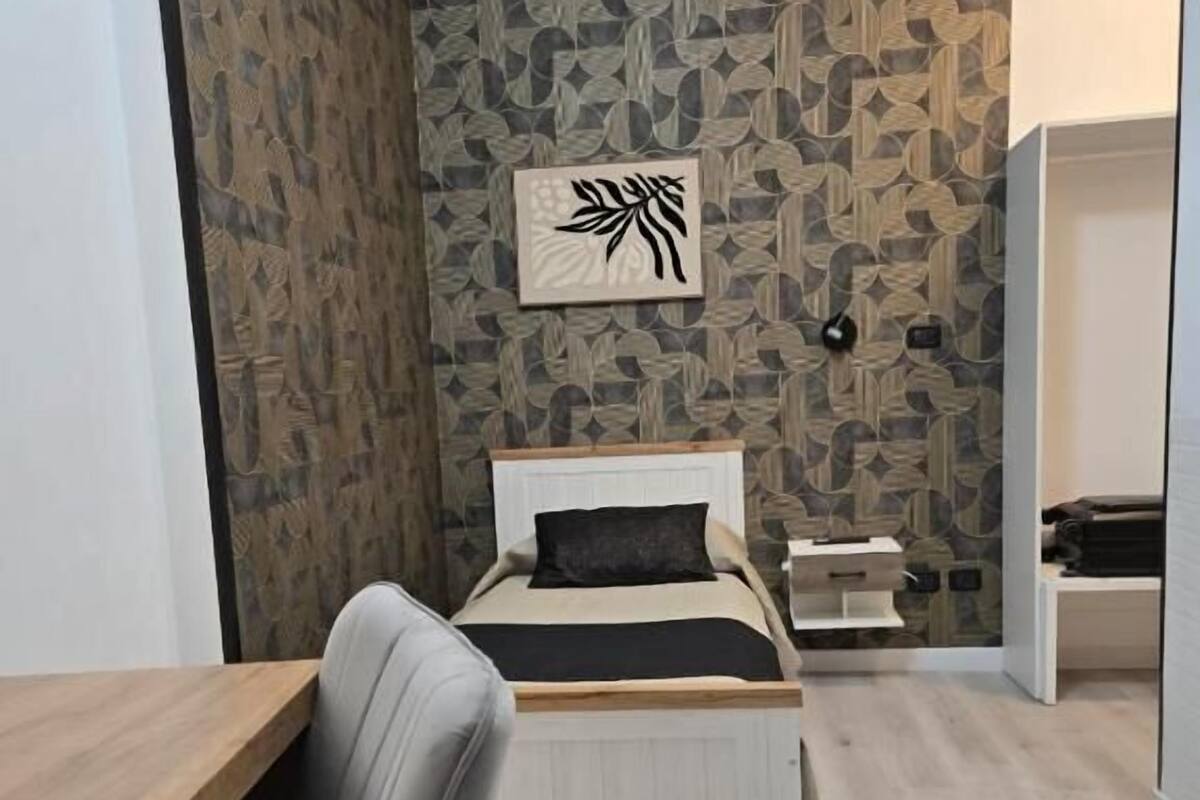 A modern bedroom with a bed, a desk, and a chair. There is a wall with a leaf pattern and a framed picture above the bed.