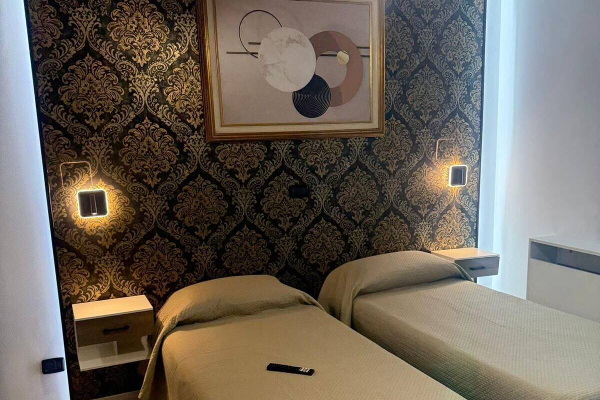 A hotel room with two beds, a patterned wallpaper, a framed artwork above the beds, and bedside lamps.