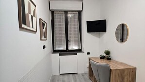 Ein kleiner, moderner Raum mit einem Holztisch, einem grauen Stuhl, einem an der Wand montierten Fernseher und einem Fenster mit Vorhängen.