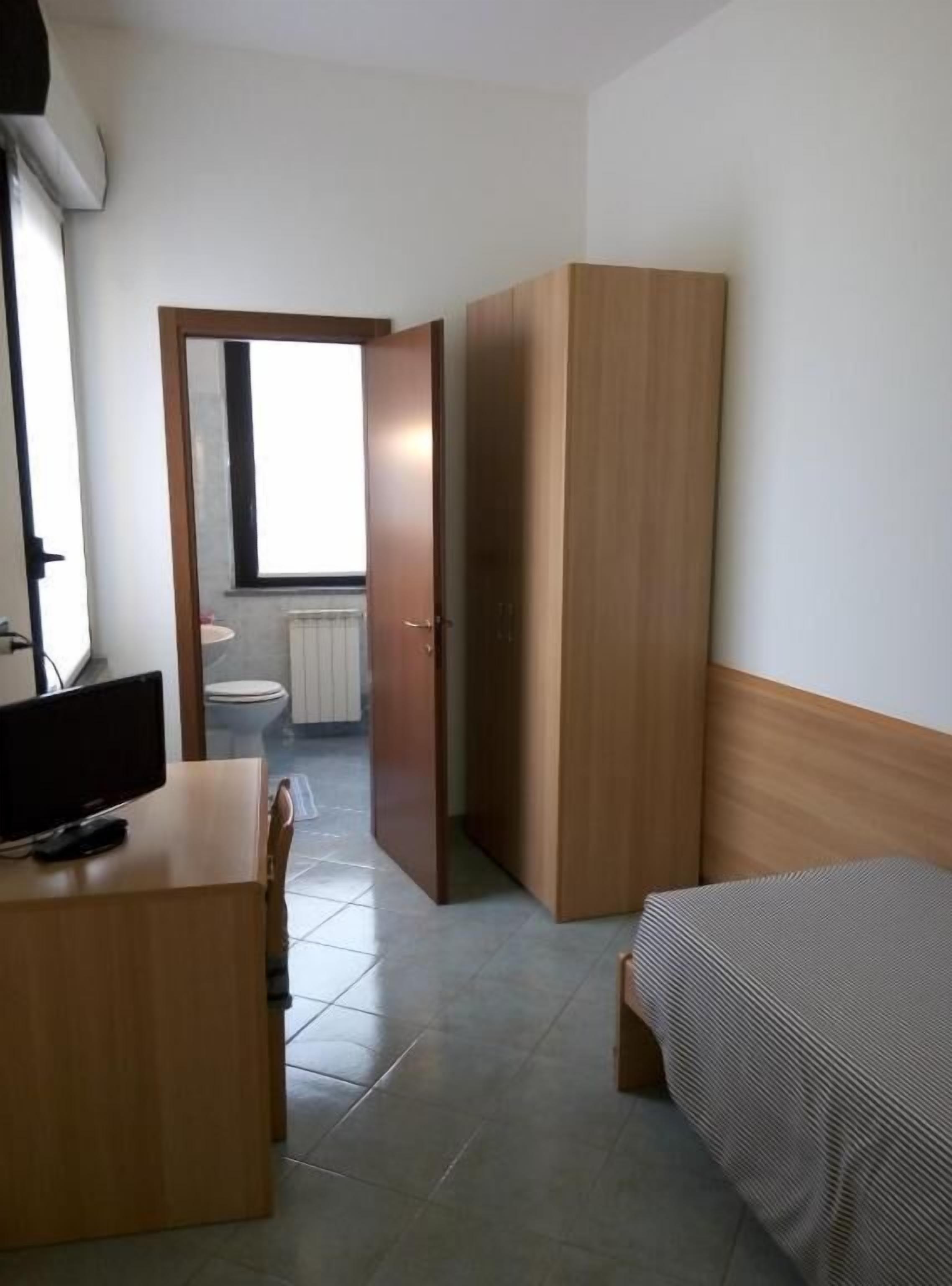 A small, modern room with a bed, a desk, and a wardrobe. There is a window providing natural light, and a toilet visible through an open door.