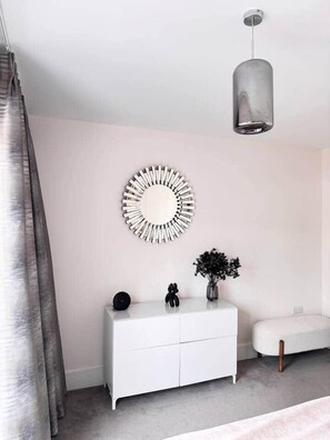Une chambre à coucher moderne avec une commode blanche, un miroir rond, un luminaire suspendu et un banc gris.