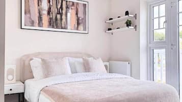 Un dormitorio con cama, mesita de noche, radiador, ventana y estante en la pared con una planta.