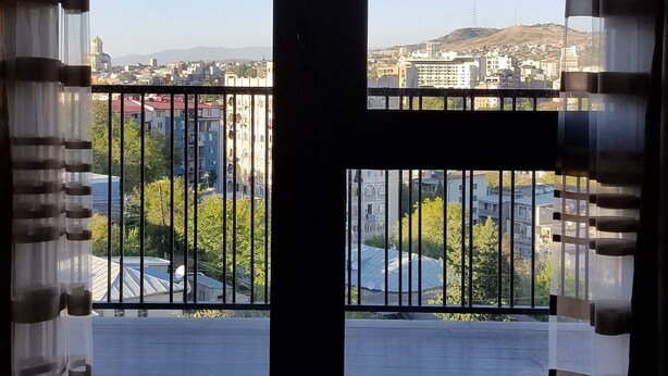 Ein Zimmer mit großem Fenster, einem Balkon mit Metallgeländer und Blick auf eine Stadtlandschaft.