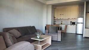 Ein modernes Wohnzimmer mit Sofa, Couchtisch und Essbereich mit Stühlen. Im Hintergrund ist eine Küche mit Schränken und Geräten zu sehen.