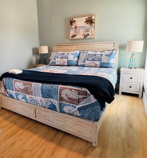 Ein Bett mit blau-weißer Bettdecke, zwei passende Kissen, ein hölzernes Kopfteil, ein an der Wand befestigtes Bild von einer Strandansicht und zwei Nachttische mit Lampen.