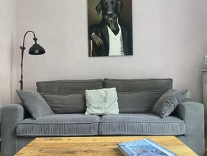 Ein graues Cordsofa mit einem weißen Kissen und zwei grauen Kissen, ein Couchtisch aus Holz mit einem Buch, eine schwarze Stehlampe und ein gerahmtes Bild von einem Hund an der Wand.