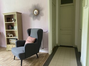 Ein Zimmer mit einem dunkelgrauen Sessel, einem rosafarbenen Kissen, einem hölzernen Bücherregal mit Büchern, einem dekorativen Spiegel und einer weißen Tür.
