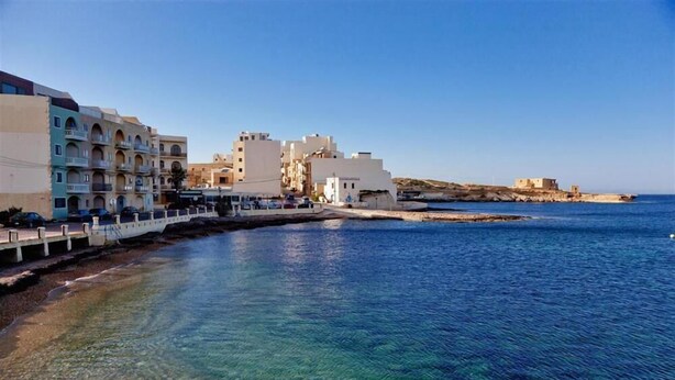 A coastal town with a clear blue sea, buildings with balconies, and a pier extending into the water. - Sea view two bedroom apartment (Marsalforn)
