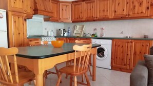 A kitchen with wooden cabinets, a dining table with chairs, a washing machine, and a grey sofa in the living area. - Sea view two bedroom apartment (Marsalforn)