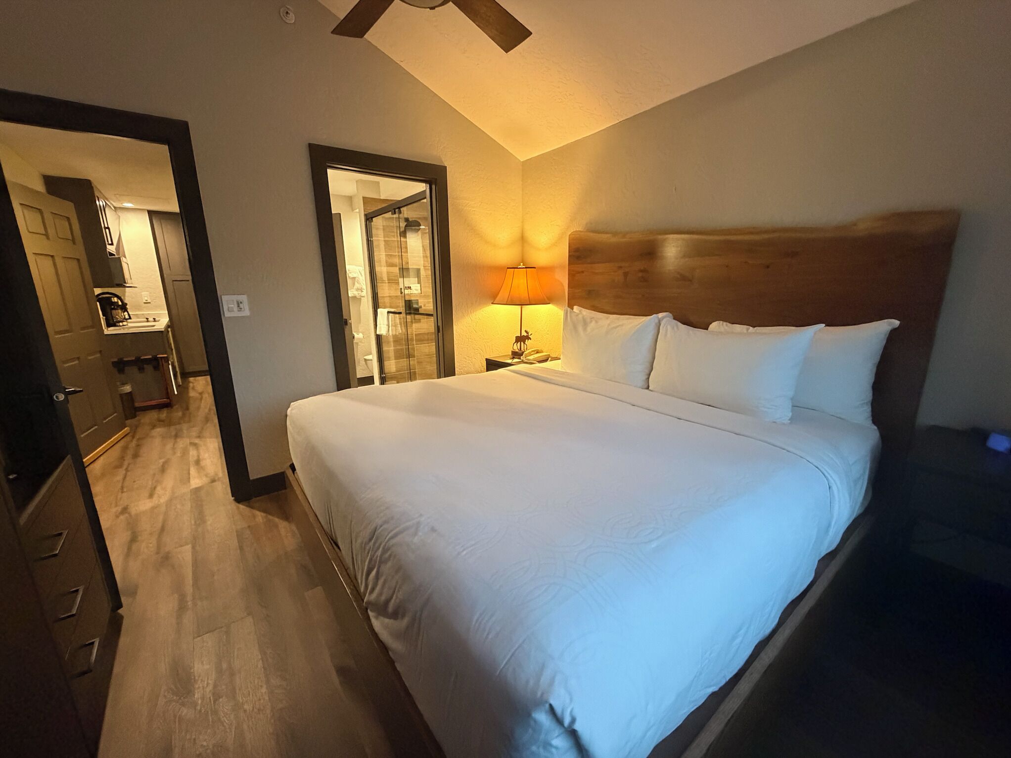 A hotel room with a large bed, wooden headboard, a bedside lamp, and a view of the bathroom and kitchen through an open door.