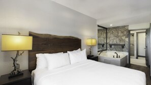 A hotel room with a large bed, two bedside lamps, a stone wall, a bathroom with a bathtub, and a door leading to another room. - Park City, Ski-in, Ski-out, 2-bed 2 bath, Sleeps 8 (Park City)