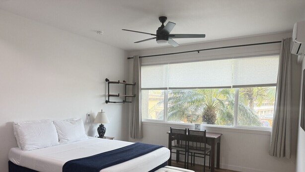 Ein Schlafzimmer mit einem Bett, einem Schreibtisch, einem Stuhl, einem Deckenventilator und einem Fenster mit Vorhängen.