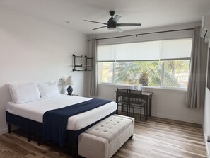 Un dormitorio con cama, escritorio, silla, ventilador de techo y ventana con cortinas.