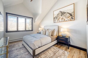 A bedroom with a large bed, two bedside tables with lamps, a window with a view, and a mountain painting on the wall. - Charlie's Slopeside: Ski-In/Out Arrowhead Penthouse, Mountain Views, Vaulted Ceilings + Balcony (Edwards)