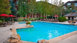 A pool area with lounge chairs and red umbrellas, surrounded by trees and a building with multiple windows. - Charlie's Slopeside: Ski-In/Out Arrowhead Penthouse, Mountain Views, Vaulted Ceilings + Balcony (Edwards)