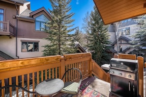 A balcony with a grill, a table, and chairs, overlooking a residential area with houses and trees. - Charlie's Slopeside: Ski-In/Out Arrowhead Penthouse, Mountain Views, Vaulted Ceilings + Balcony (Edwards)