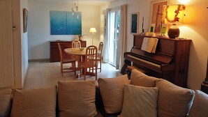 Ein Wohnzimmer mit einem Klavier, einem Esstisch mit Stühlen, einem Sofa mit Kissen, einer Lampe und einem Gemälde an der Wand.