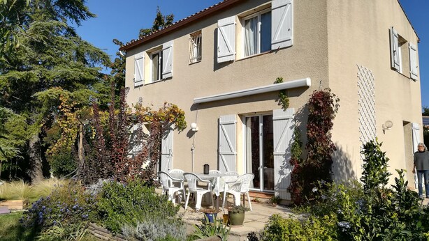 Ein zweistöckiges Haus mit Garten, Sitzgelegenheiten im Freien und einer Tür, die auf eine überdachte Terrasse führt.