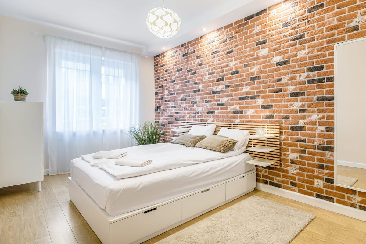 A bedroom with a brick wall, a bed, a bedside table, and a mirror.