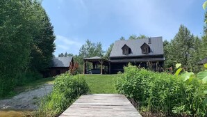 Eine Holzterrasse führt zu einem Haus mit Metalldach, umgeben von üppigem Grün und blauem Himmel.