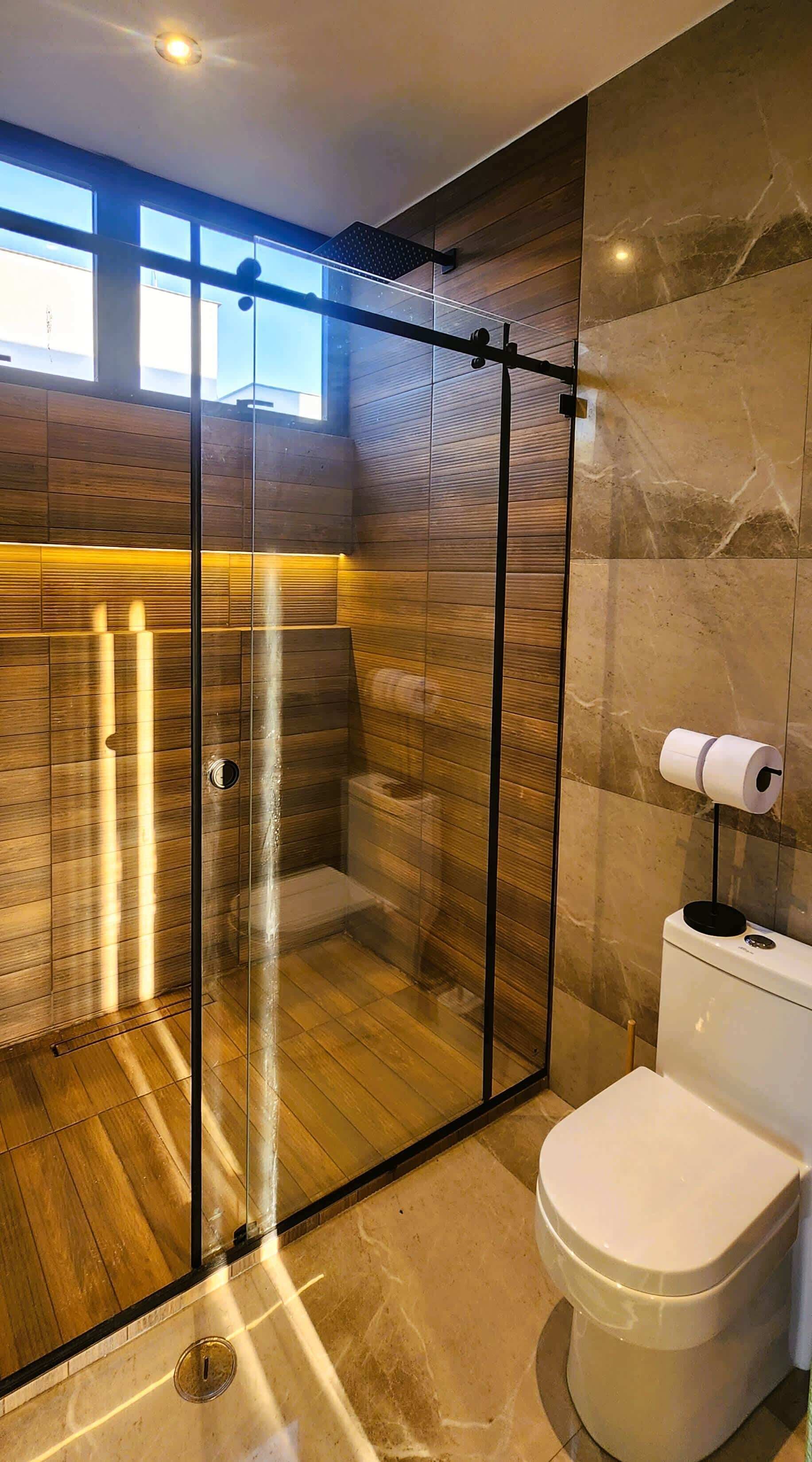 Ein modernes Badezimmer mit einer gläsernen Duschkabine, Holzpaneelen und einem WC mit Papierhalter.