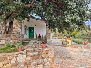 A stone-walled building with a green door, a wooden table and chairs outside, and a garden with potted plants.