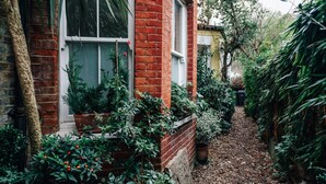En smal brobelagt sti kantet med frodigt grønt og plantekasser, der fører hen til et hus med en rød murstensfacade og vinduer med hvide rammer.