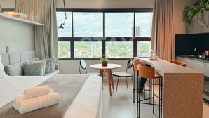 Une chambre d’hôtel moderne comprenant un lit, un coin repas, une cuisine et une grande fenêtre donnant sur la ville.