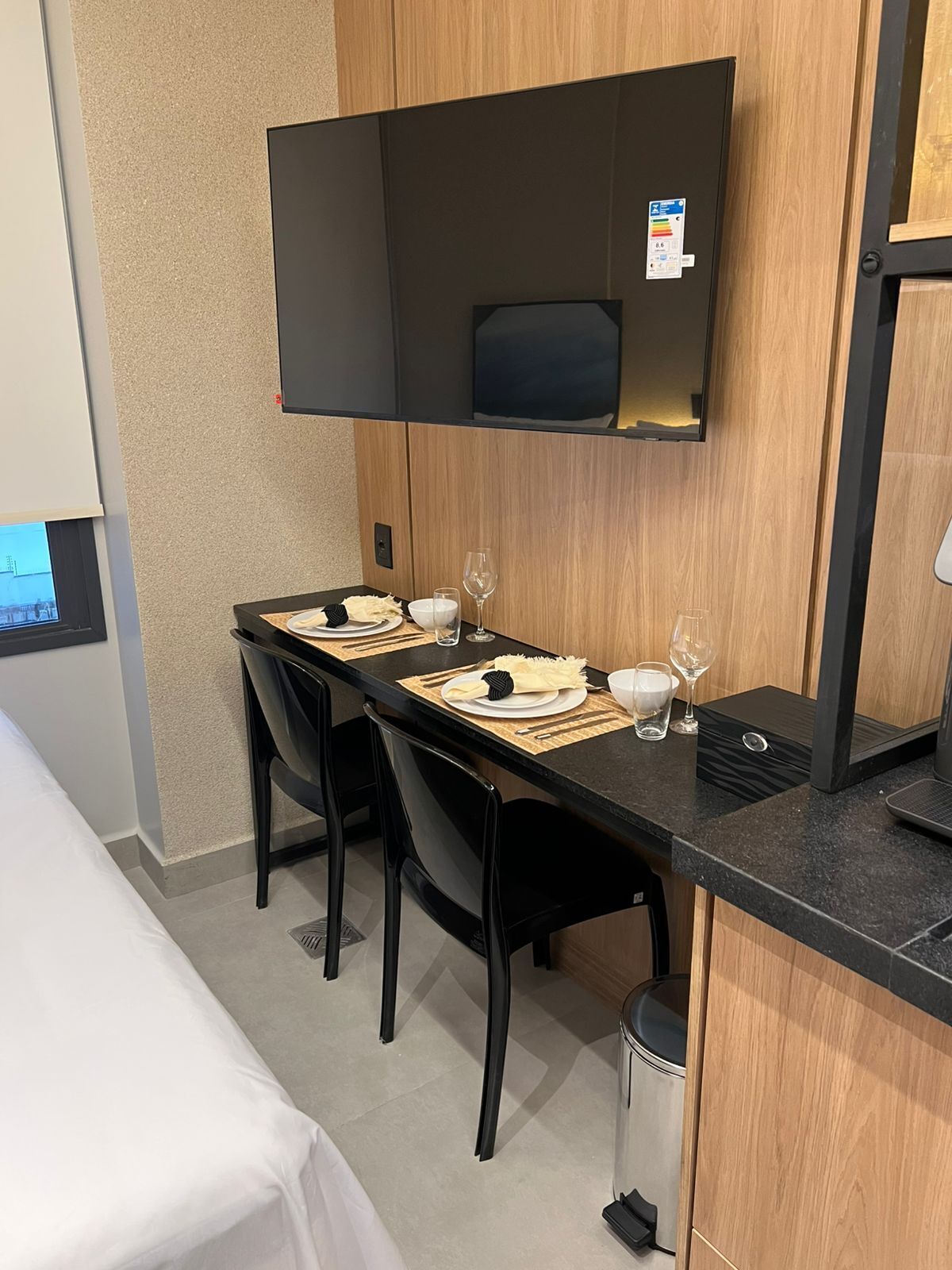 A hotel room with a dining table set for two, a TV mounted on the wall, and a bed with a white cover.