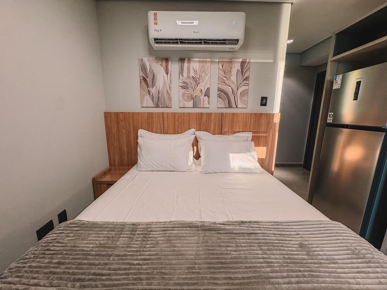 A hotel room with a bed, nightstand, wall-mounted air conditioning unit, and a refrigerator.
