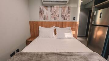A hotel room with a bed, nightstand, wall-mounted air conditioning unit, and a refrigerator.