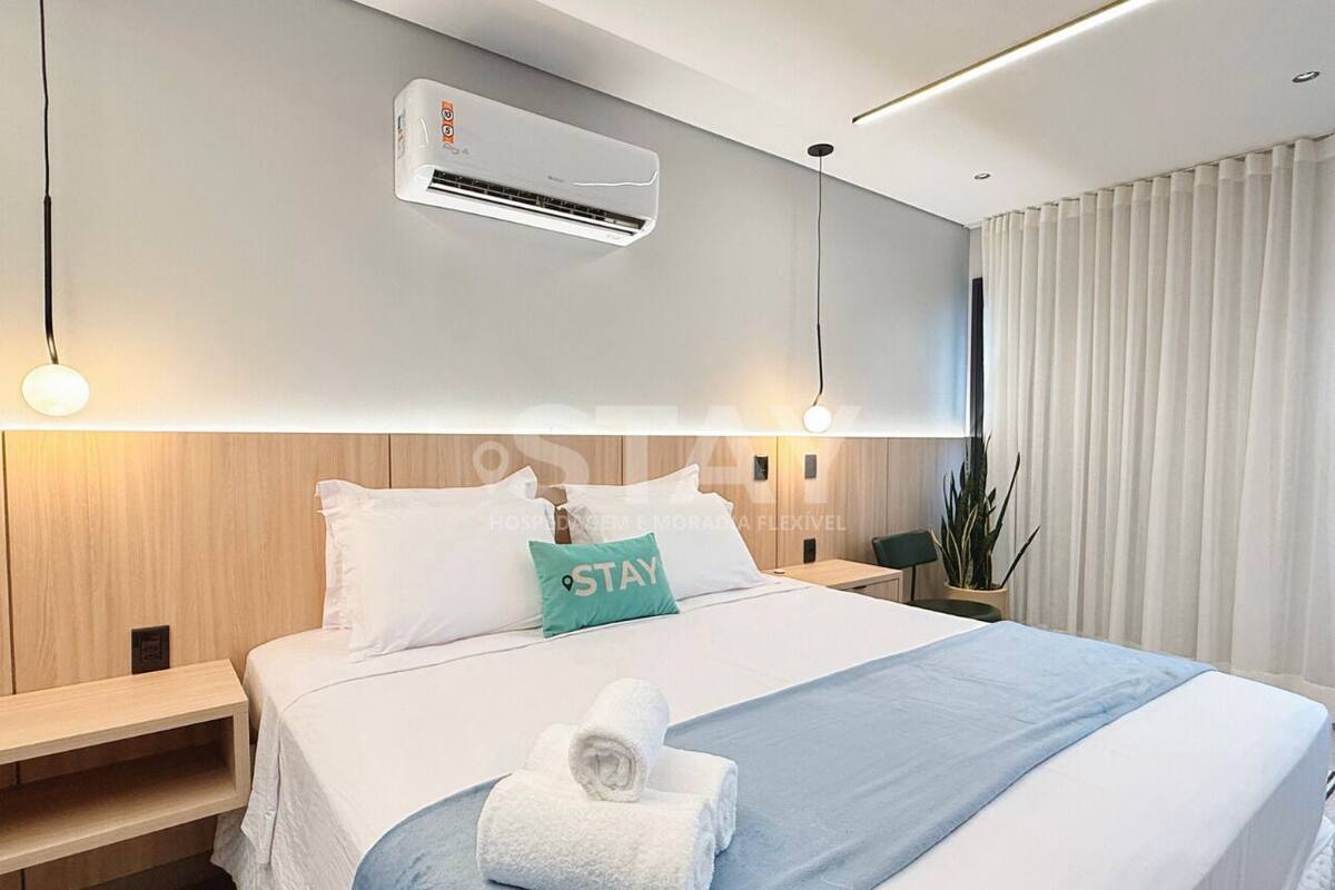 A hotel room with a bed, a nightstand, a wall-mounted air conditioner, a potted plant, and a window with curtains.