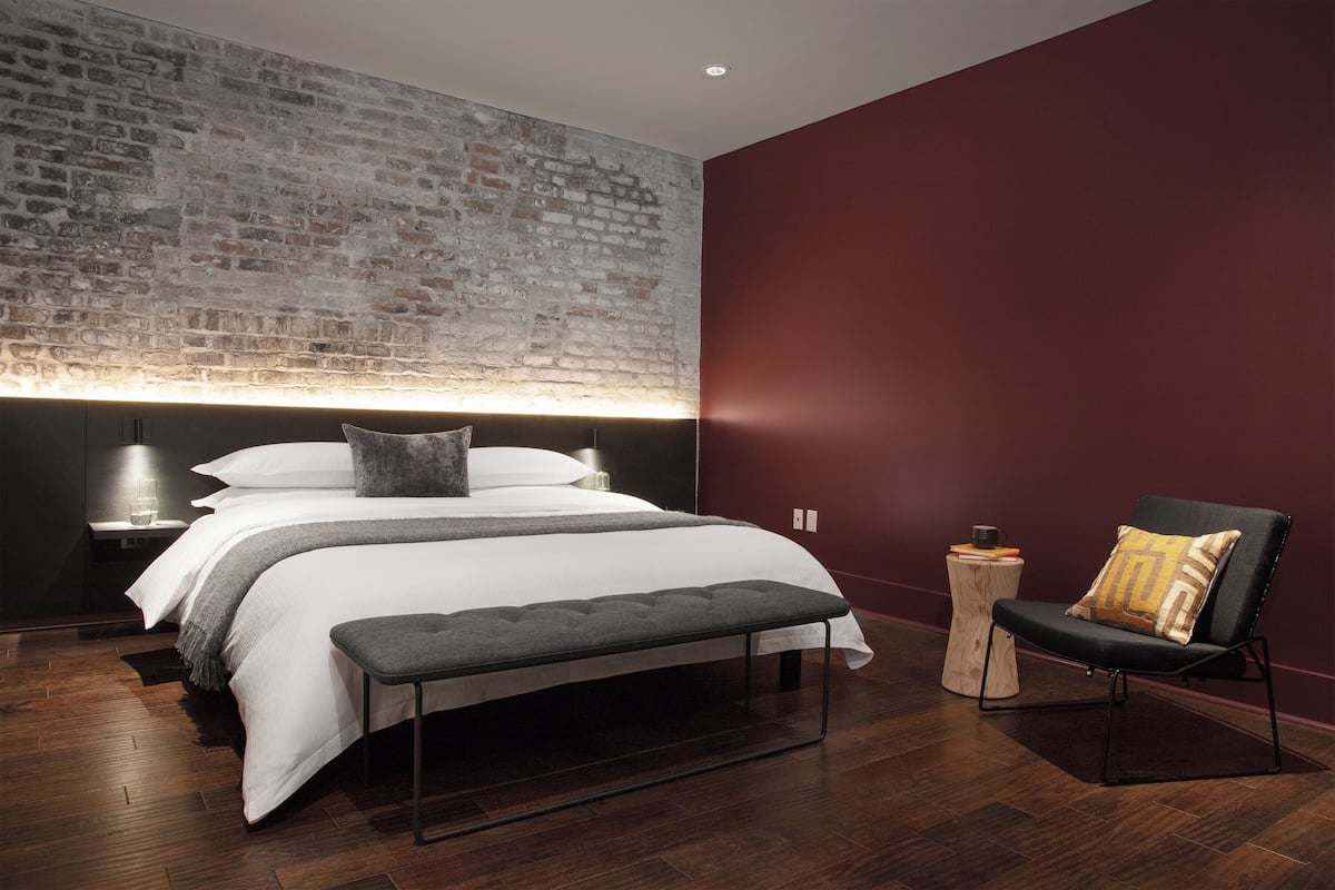 A modern bedroom with a brick wall, a large bed, a bench, a chair, and a nightstand with a lamp.