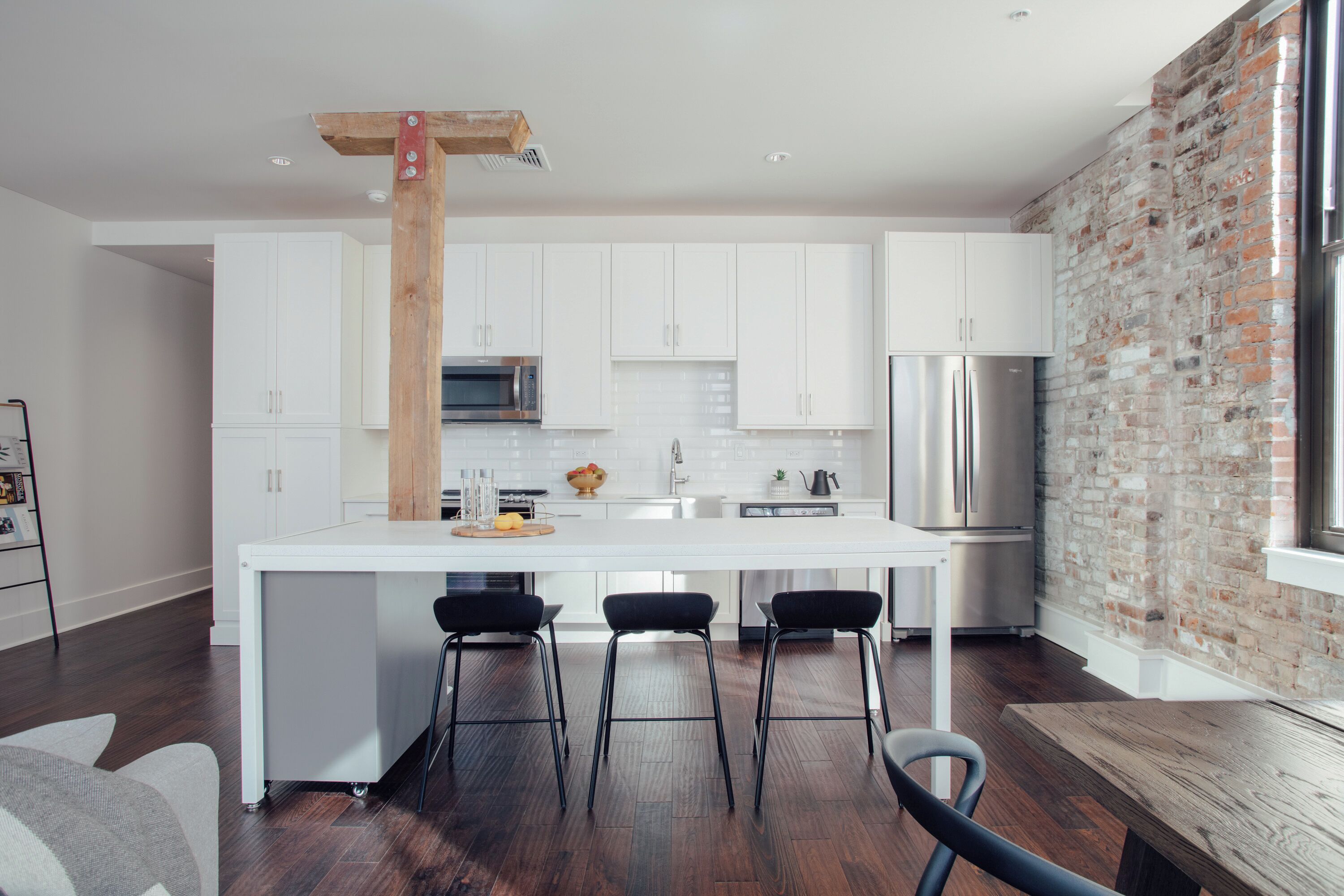 Una cocina moderna con gabinetes blancos, una isla central con taburetes, una pared de acento de ladrillo y un refrigerador de acero inoxidable.